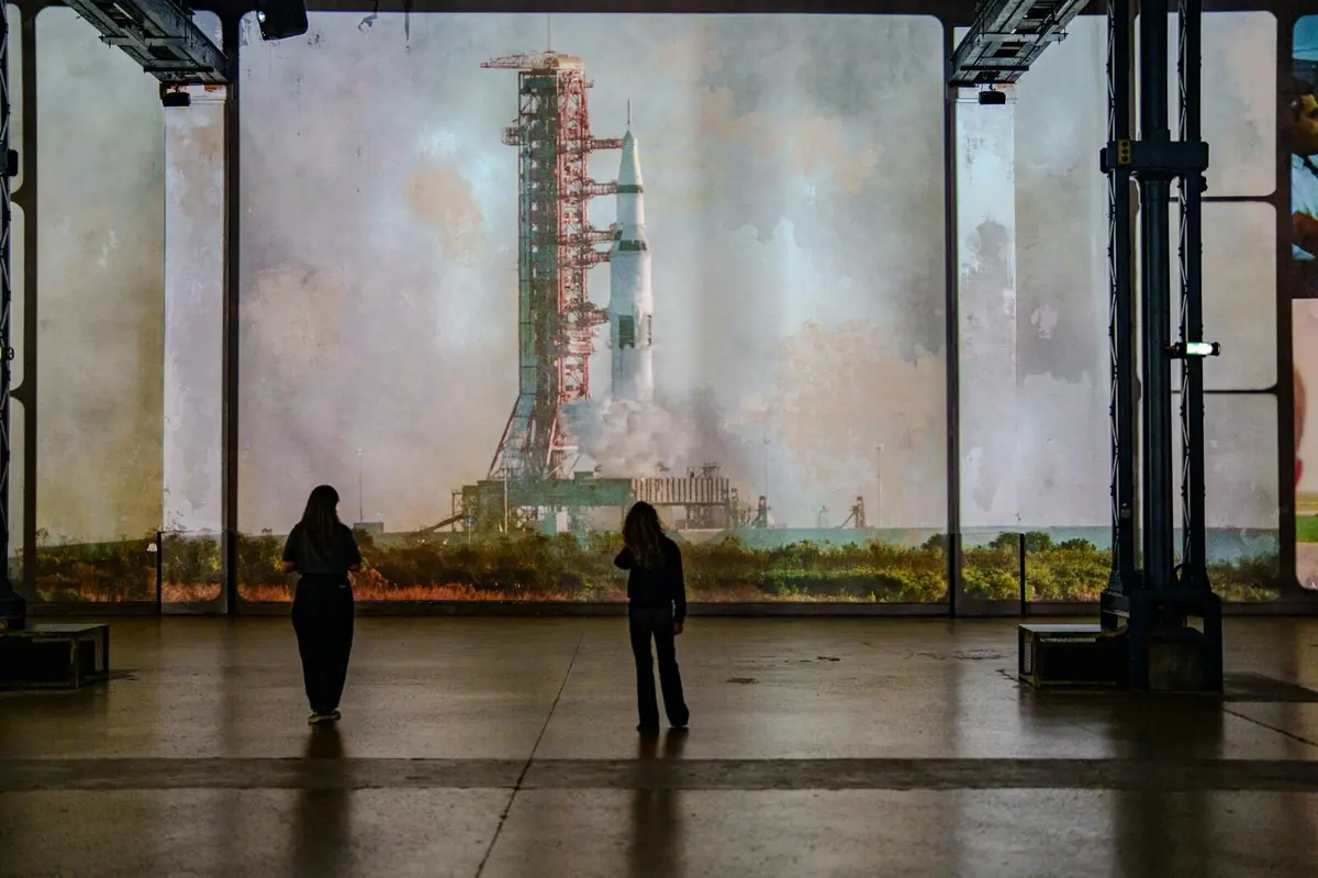 Deux personnes se tiennent à l'intérieur, face à un grand écran montrant une fusée sur une rampe de lancement avec de la fumée à sa base.