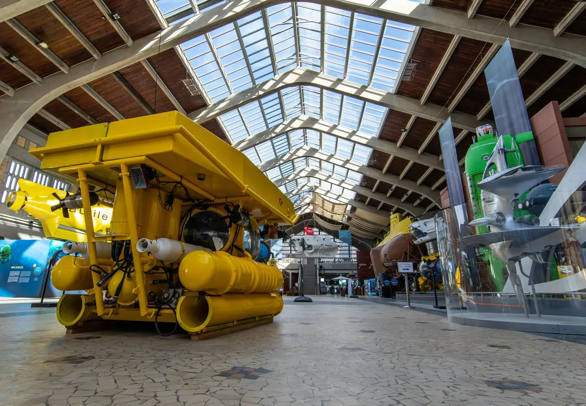 De grands submersibles jaunes sont exposés à l'intérieur d'un bâtiment au haut plafond vitré et au sol en dalles de pierre.