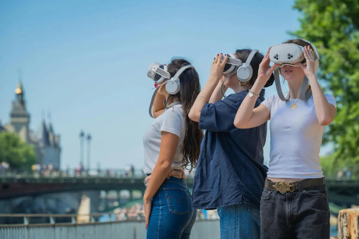 Trois personnes portant des casques de réalité virtuelle à l'extérieur, près d'un pont, par une journée ensoleillée.
