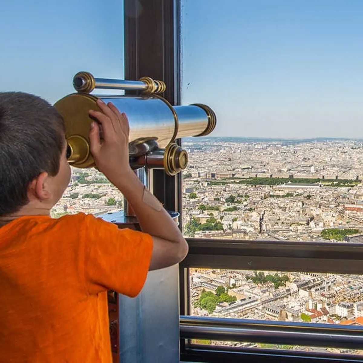 Un garçon vêtu d'une chemise orange regarde dans un télescope un paysage urbain depuis un point d'observation élevé par temps clair.