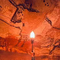 Une main tenant une torche éclaire des peintures rupestres préhistoriques d'animaux sur une surface rocheuse texturée. DR