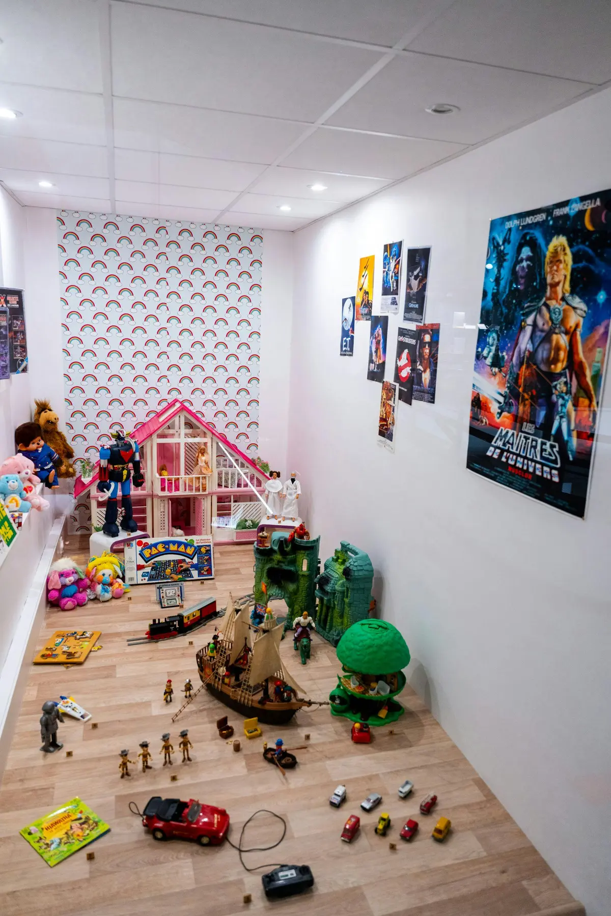 Une salle de jeux pour enfants avec des jouets, une maison de poupée, des figurines et des posters sur les murs. Le sol est parsemé de divers jeux.
