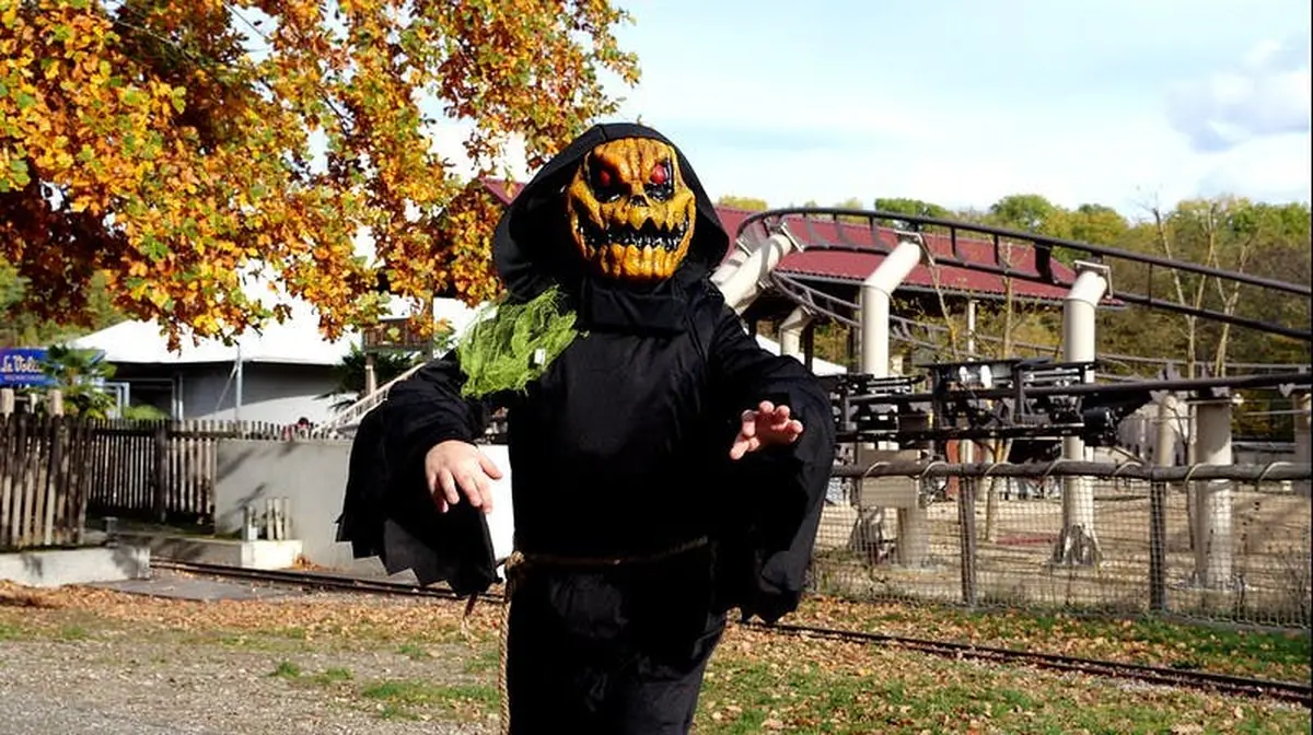 Personne portant un costume noir à capuche et un masque de citrouille posant dans un parc avec des feuilles d'automne et une structure métallique en arrière-plan.