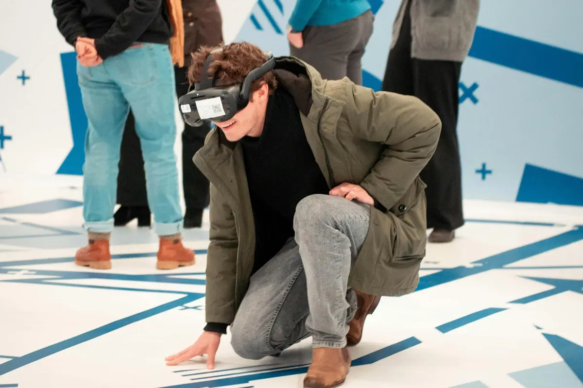 Une personne portant des lunettes de réalité virtuelle est accroupie sur un sol à motifs géométriques, tandis que d'autres personnes se tiennent à l'arrière-plan.
