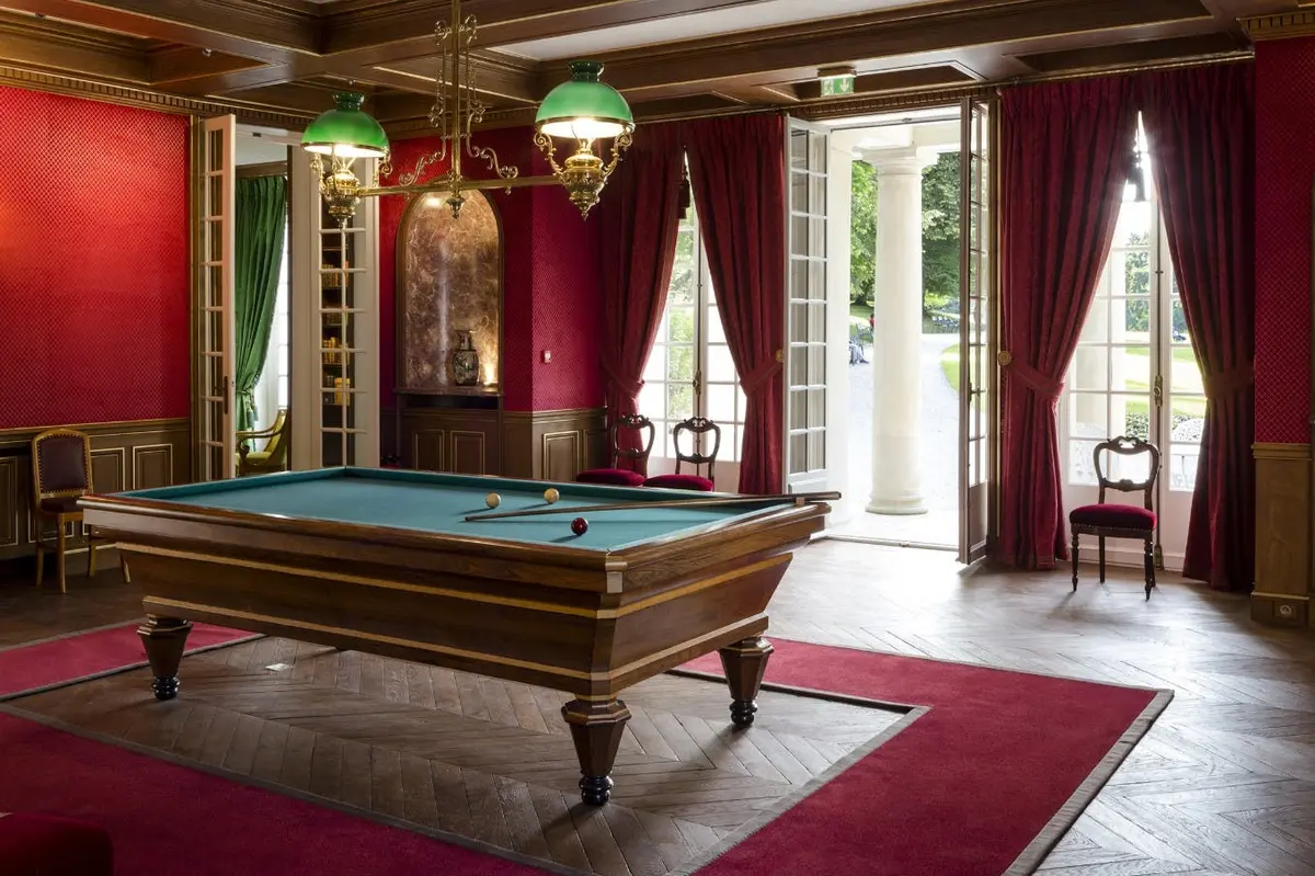 Une table de billard dans une pièce luxueuse avec des rideaux rouges, des meubles en bois et des éclairages décoratifs. Des portes ouvertes donnent sur un jardin.