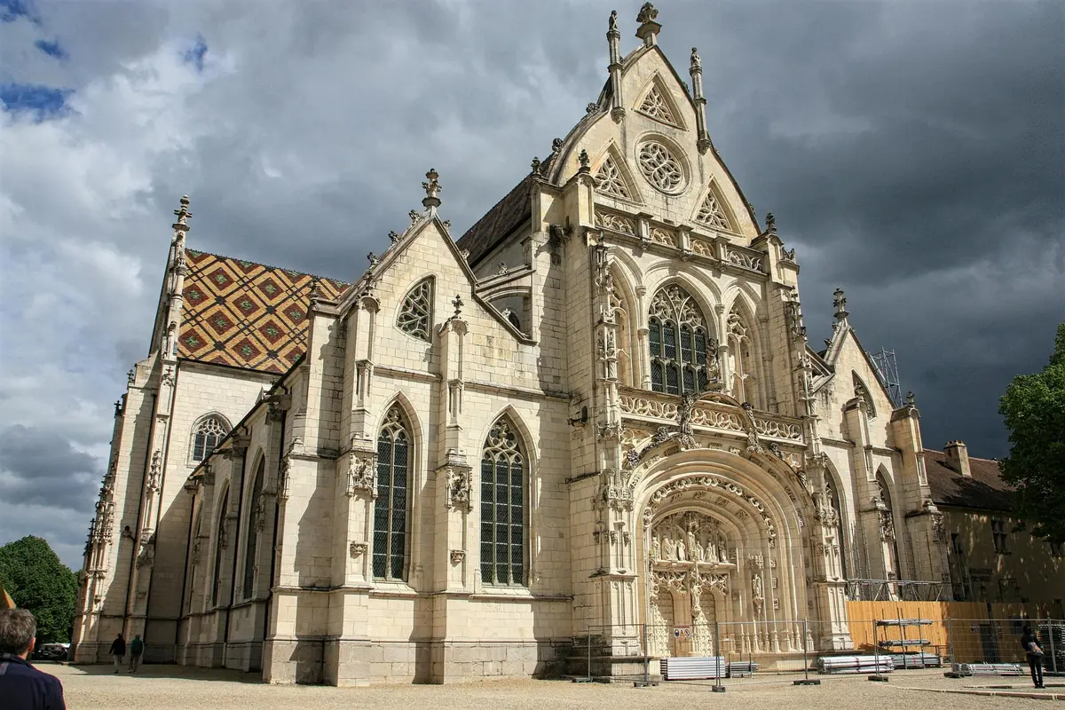 Cathédrale de style gothique avec une façade en pierre ornée, des fenêtres cintrées et des sculptures détaillées sur fond de ciel nuageux.