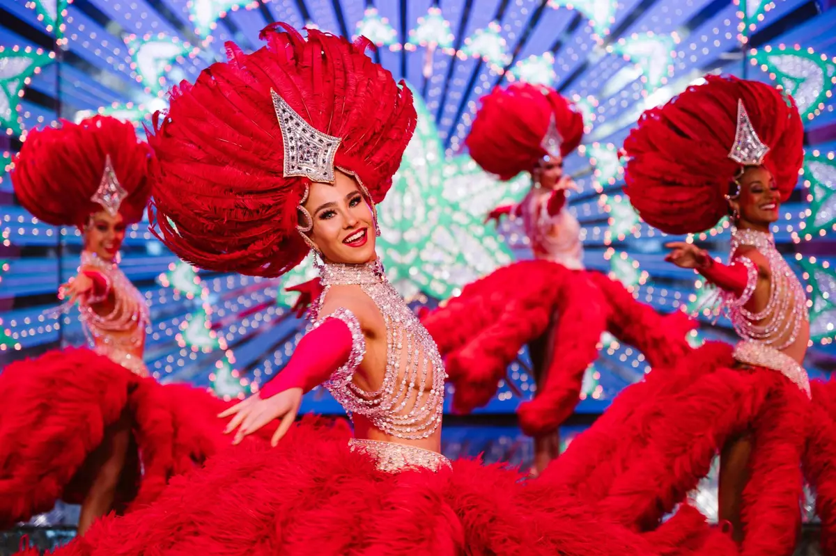 Des artistes vêtus de costumes rouges somptueux et coiffés de plumes dansent sur une scène brillamment éclairée.