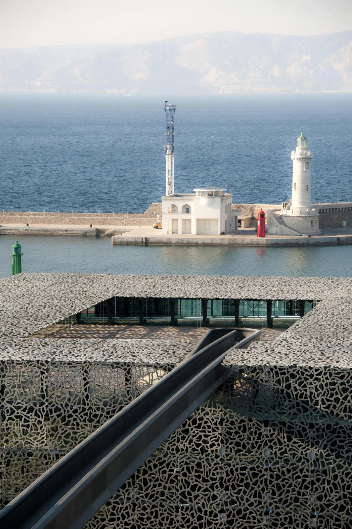 Un bâtiment moderne avec un toit à motifs surplombant l'eau, avec en arrière-plan un phare et un petit bâtiment blanc sur un brise-lames.