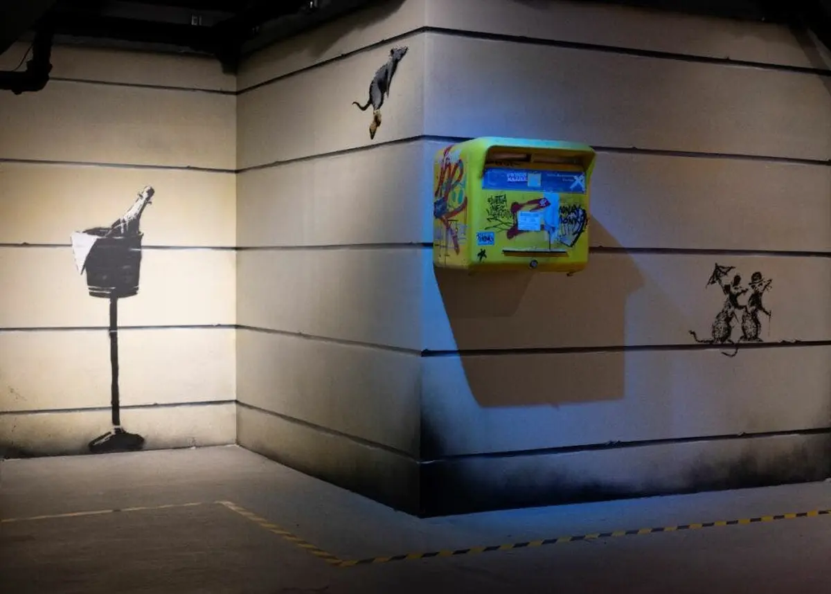 Coin d'une pièce avec art mural, représentant un homme versant un liquide d'une bouteille et un rat. Une boîte aux lettres jaune est fixée au mur.