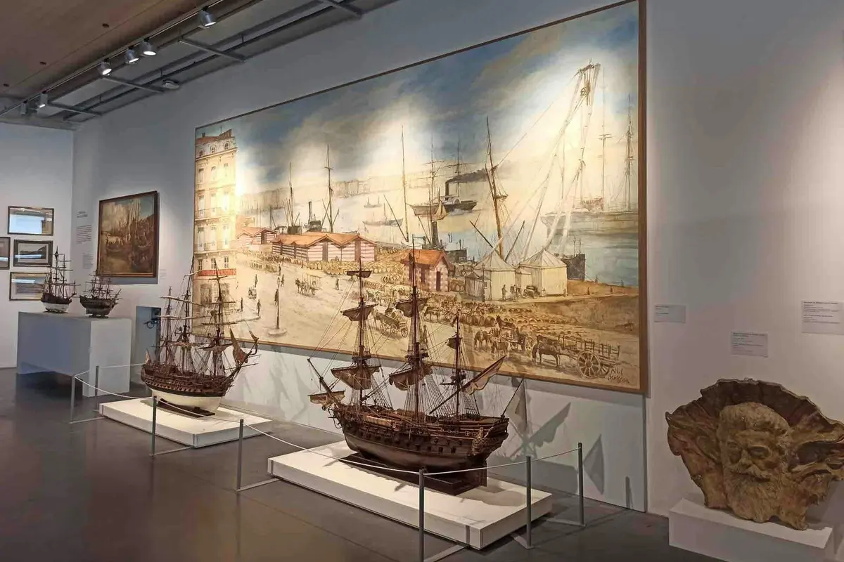 Une exposition de musée présentant une grande peinture maritime d'une scène portuaire et des modèles de navires détaillés exposés devant elle.