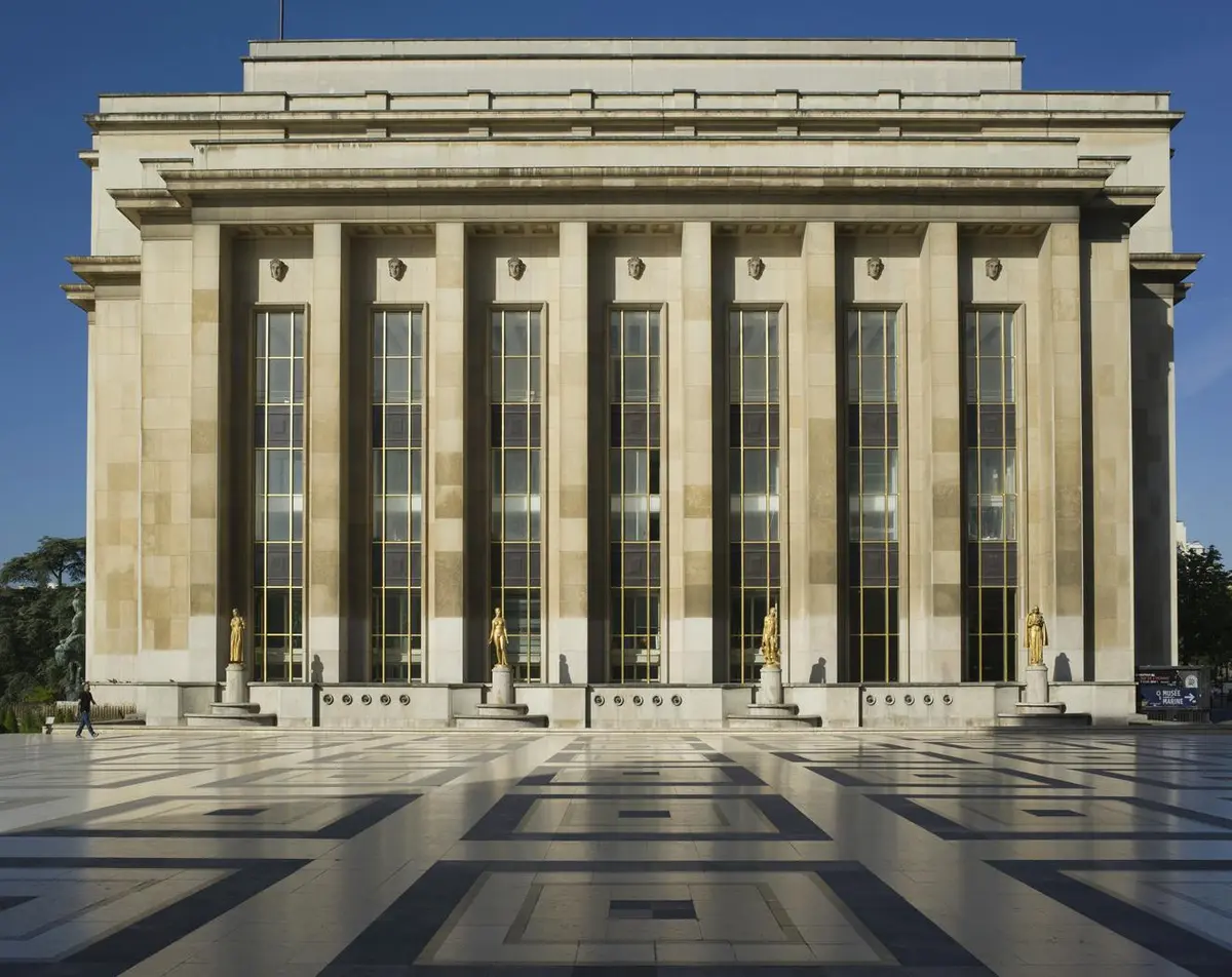 Façade d'un grand bâtiment avec de hautes colonnes, de grandes fenêtres et des statues sur une place en pierre à motifs sous un ciel bleu clair.
