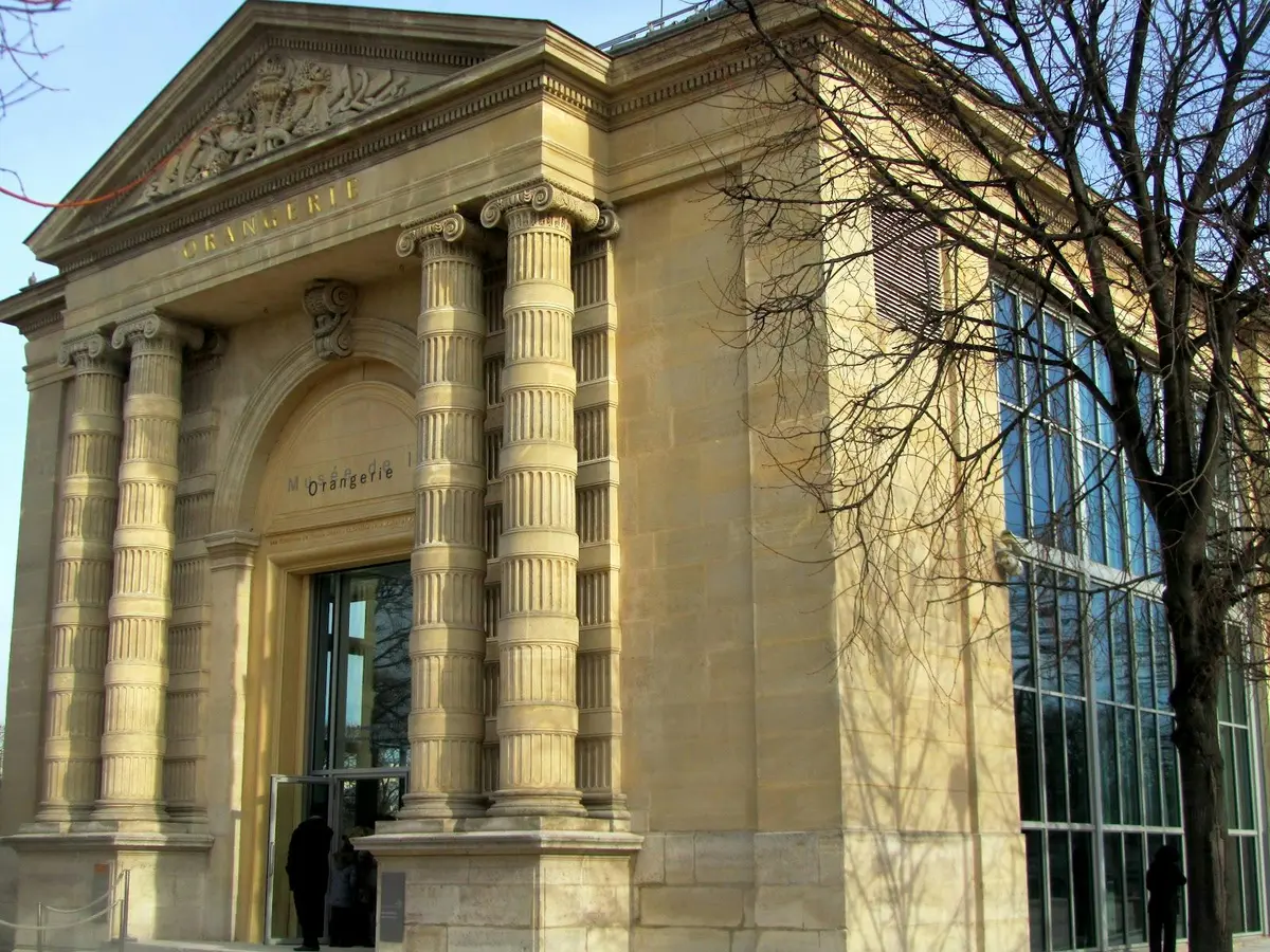 Entrée ornée d'un bâtiment historique avec de hautes colonnes cannelées et des inscriptions. Un mur de verre et un arbre se trouvent à droite.