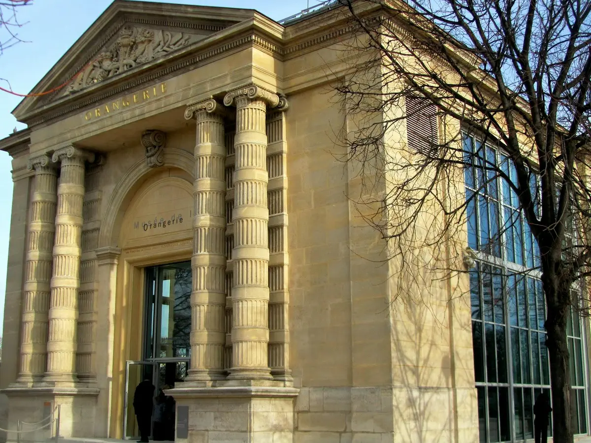 Entrée ornée d'un bâtiment historique avec de hautes colonnes cannelées et des inscriptions. Un mur de verre et un arbre se trouvent à droite.