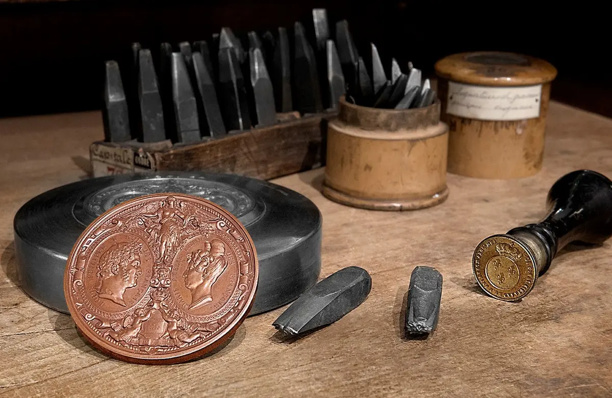 Gros plan sur des outils de gravure, une pièce de monnaie agrandie avec deux profils de face, une pièce d'or et divers burins sur une table en bois.