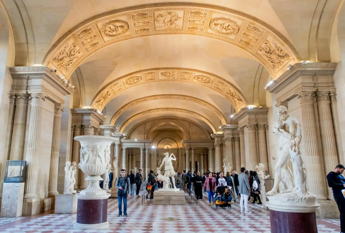 Une salle de musée avec des plafonds voûtés ornés, des sculptures et des visiteurs marchant sur un sol en damier.
