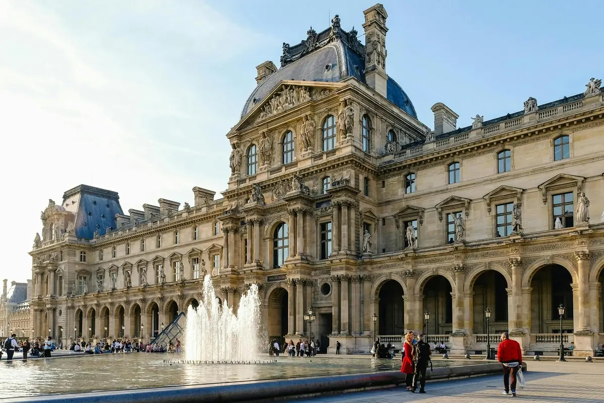 Un bâtiment historique à l'architecture ornée, une fontaine devant, et des gens qui se promènent à proximité par une journée ensoleillée.