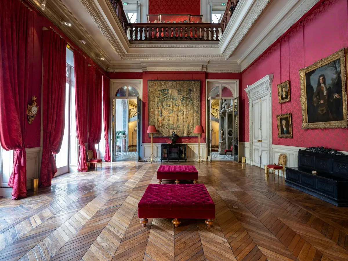 Salle élégante avec murs et rideaux rouges, parquet en bois, plafond orné, tapisserie, tableaux et sièges rouges en peluche.