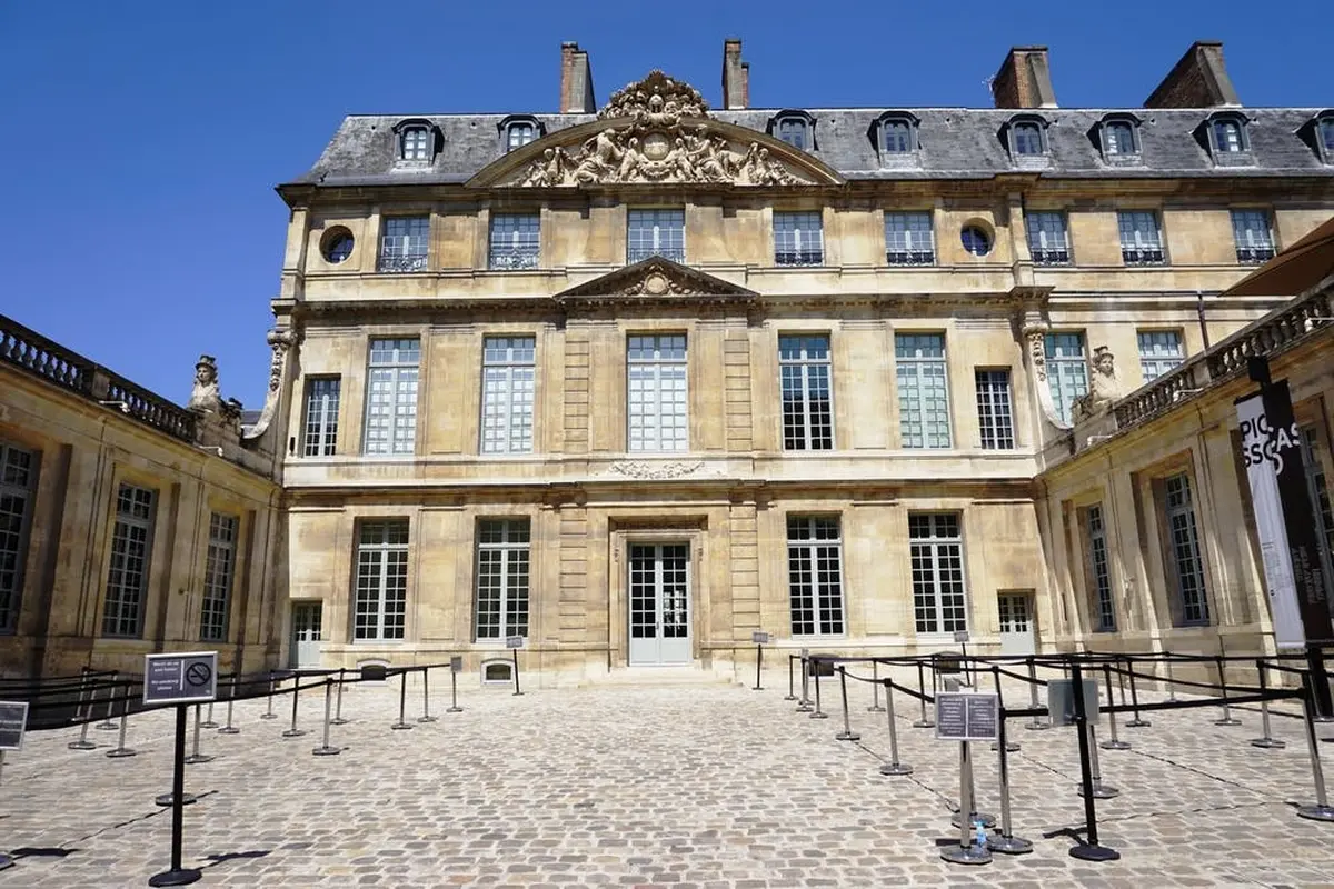 Un grand bâtiment orné de multiples fenêtres, de pierres décoratives et d'une cour pavée sous un ciel bleu clair.