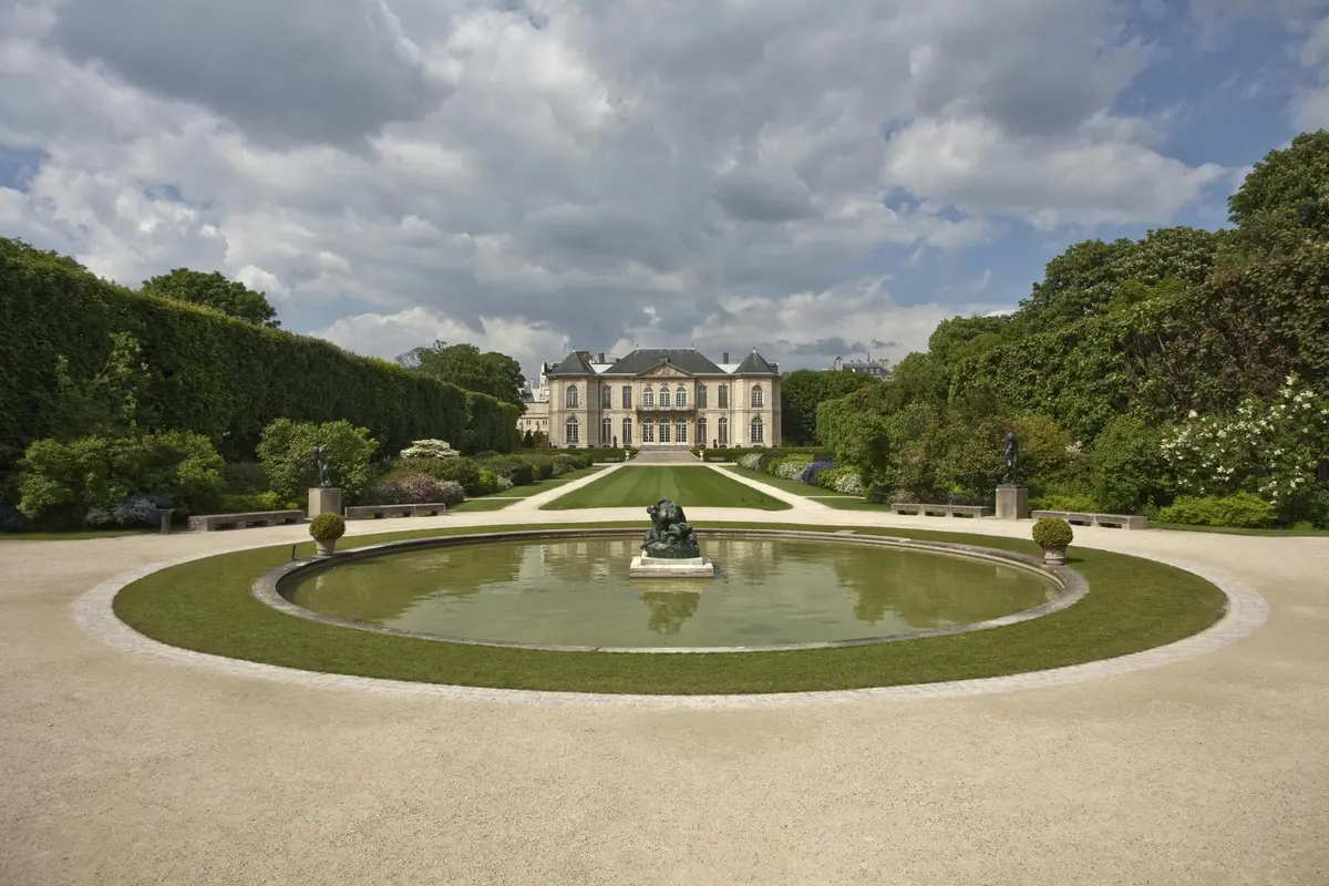 Un jardin à la française avec un étang circulaire et une statue en son centre, menant à une grande et élégante demeure sous un ciel nuageux.