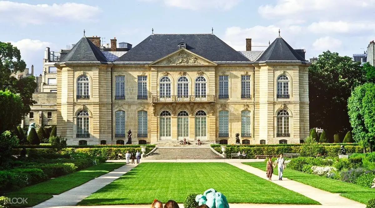Un grand bâtiment avec des fenêtres cintrées et une pelouse bien entretenue, avec une façade symétrique et plusieurs personnes dans le jardin.