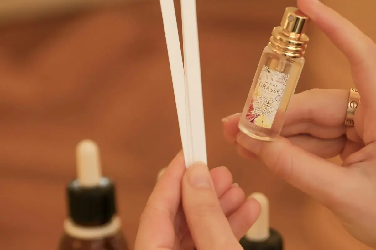 Gros plan sur des mains tenant des bandelettes de test olfactif et un flacon de parfum portant l'étiquette « Grasse », avec des flacons compte-gouttes en arrière-plan.