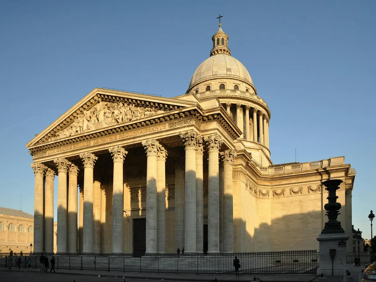 Un bâtiment néoclassique avec de hautes colonnes et un grand dôme sous un ciel bleu clair. Peu de gens se promènent près de la zone clôturée.