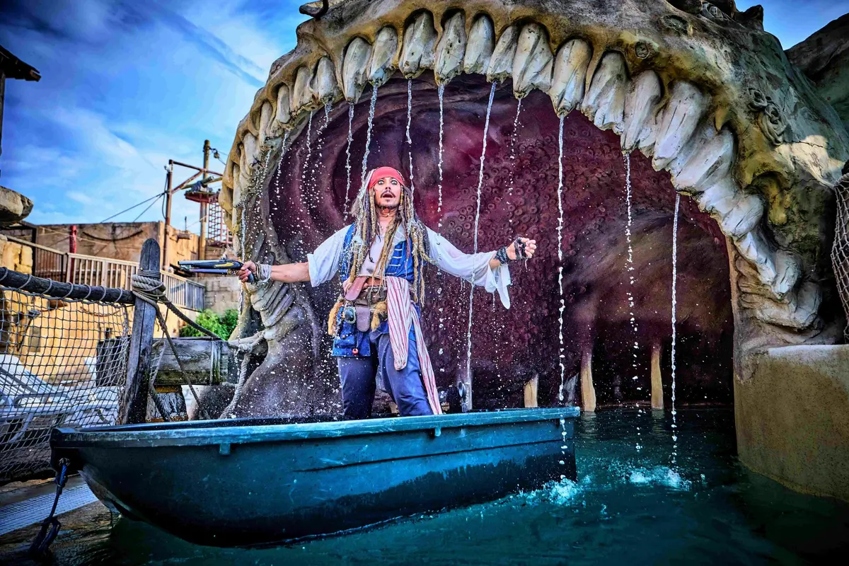 Une personne habillée en pirate se tient dans un bateau près d'une grande sculpture de baleine à la bouche ouverte, entourée d'eau.
