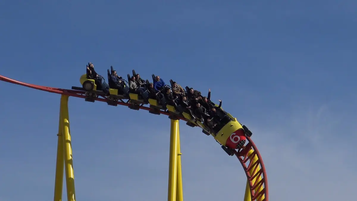 Des personnes montées sur des montagnes russes jaunes et rouges, descendant une courbe abrupte dans un ciel bleu clair.