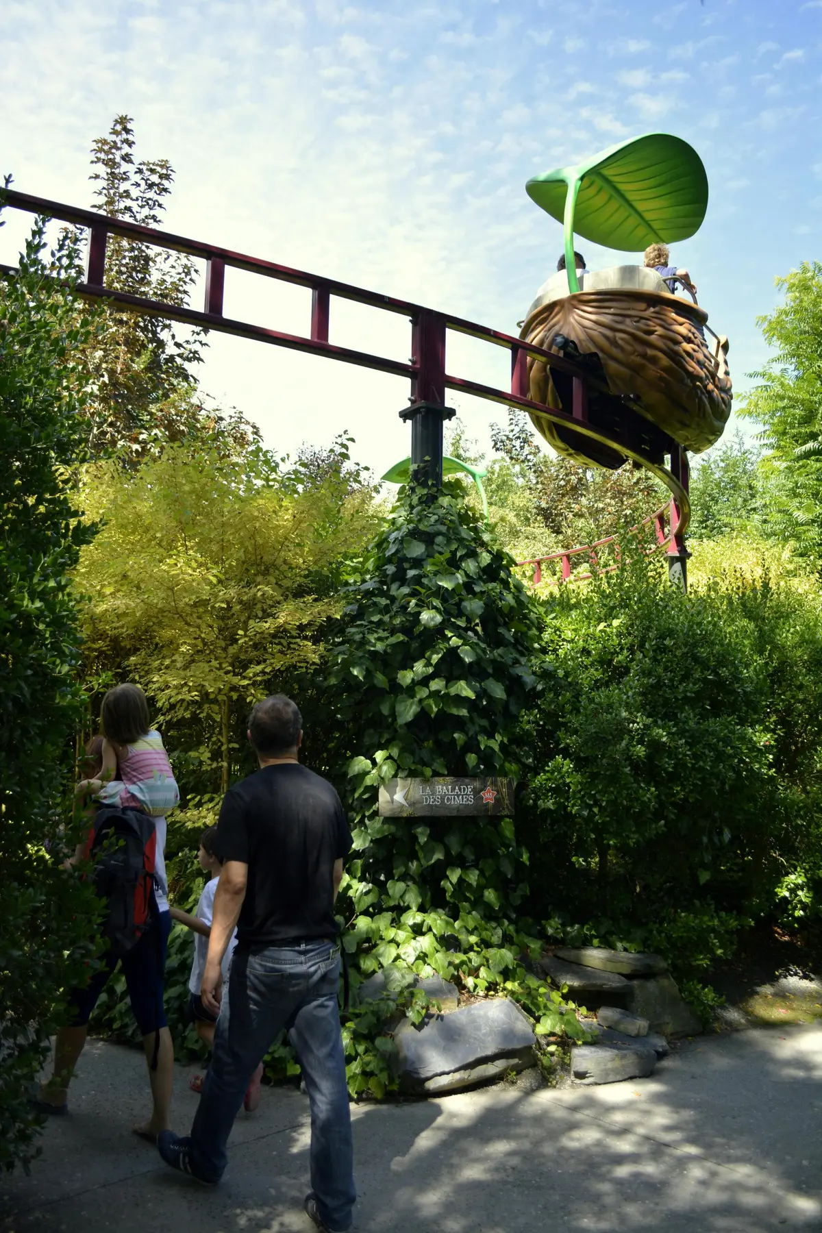 Une famille se promène sous une piste de montagnes russes rouges, entourée de verdure et d'arbres denses, avec un chariot de manège sur le thème du nid au-dessus.