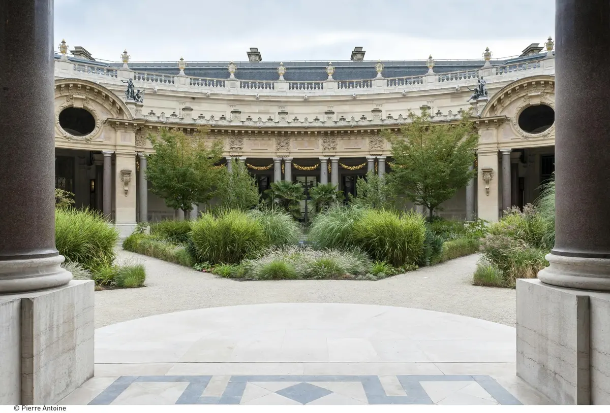 Ornate building facade with arches and columns, surrounded by lush greenery and small trees in a courtyard.