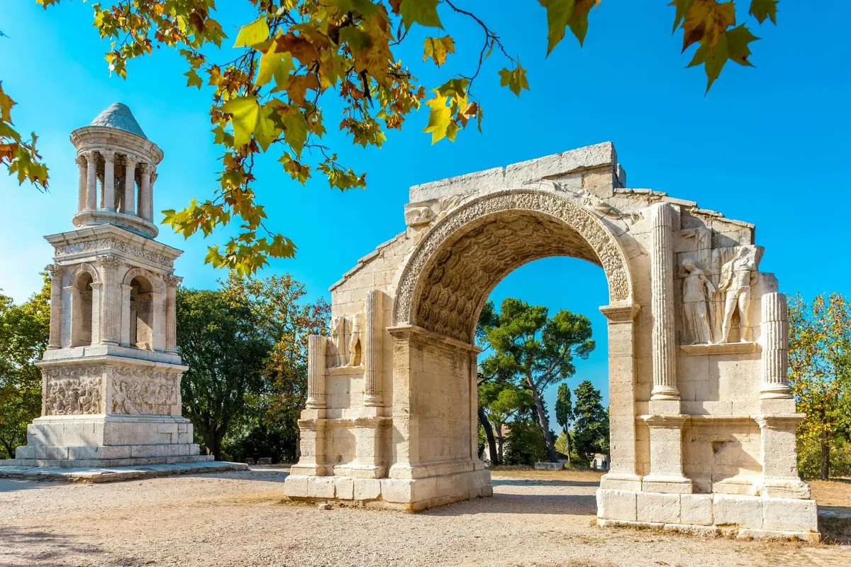 Arche et monument anciens en pierre sous un ciel bleu clair, avec des branches et des arbres feuillus en arrière-plan.