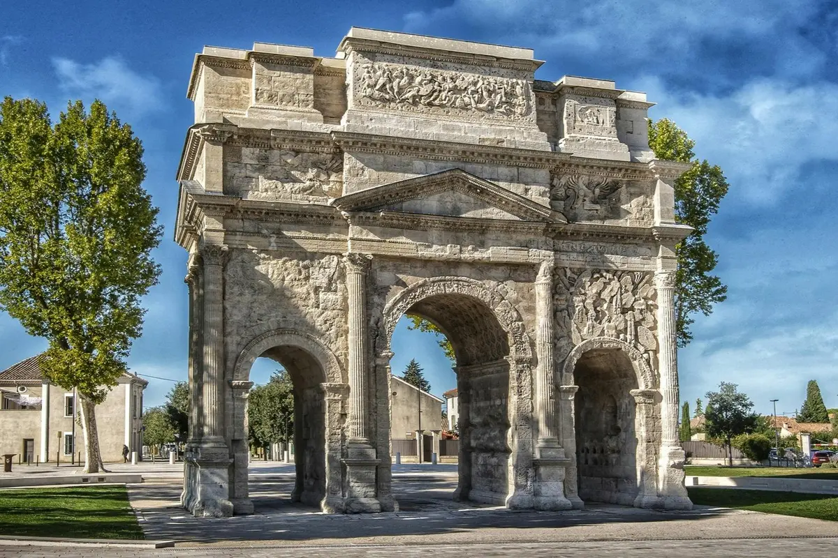 Un arc de triomphe historique en pierre, orné de sculptures détaillées et comportant trois arcades, entouré d'arbres sous un ciel bleu.