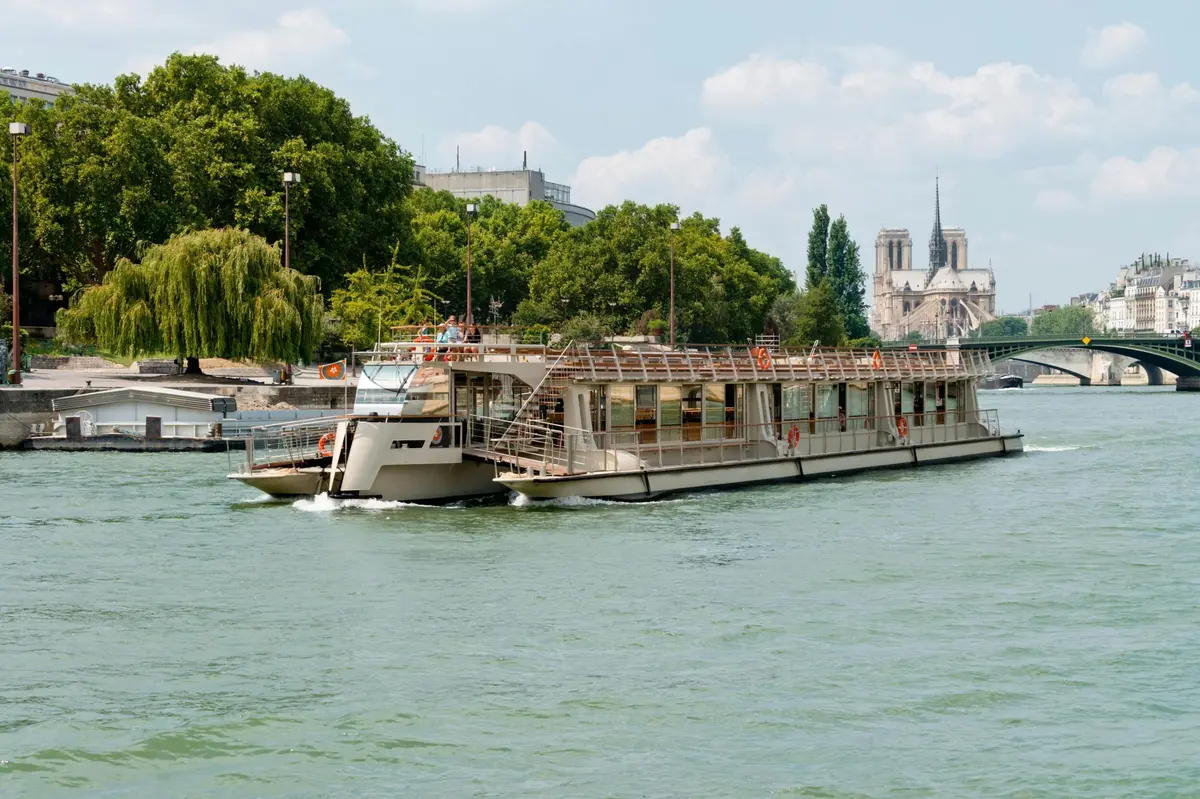 Un grand bateau d'excursion naviguant sur un fleuve, avec des arbres et une cathédrale historique en arrière-plan.