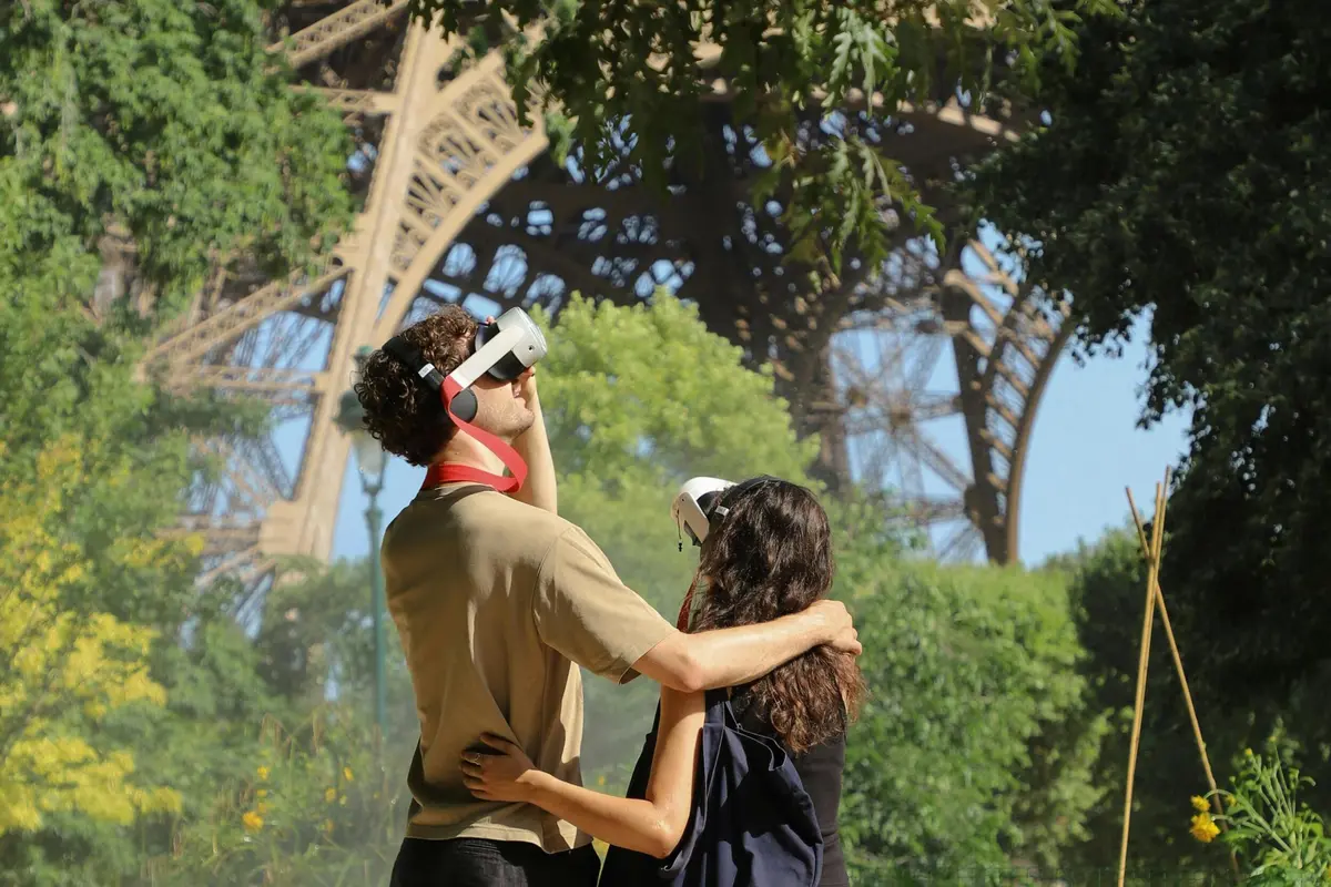Un couple portant des casques de réalité virtuelle se tient à l'extérieur près de la Tour Eiffel, entouré d'arbres et de verdure.