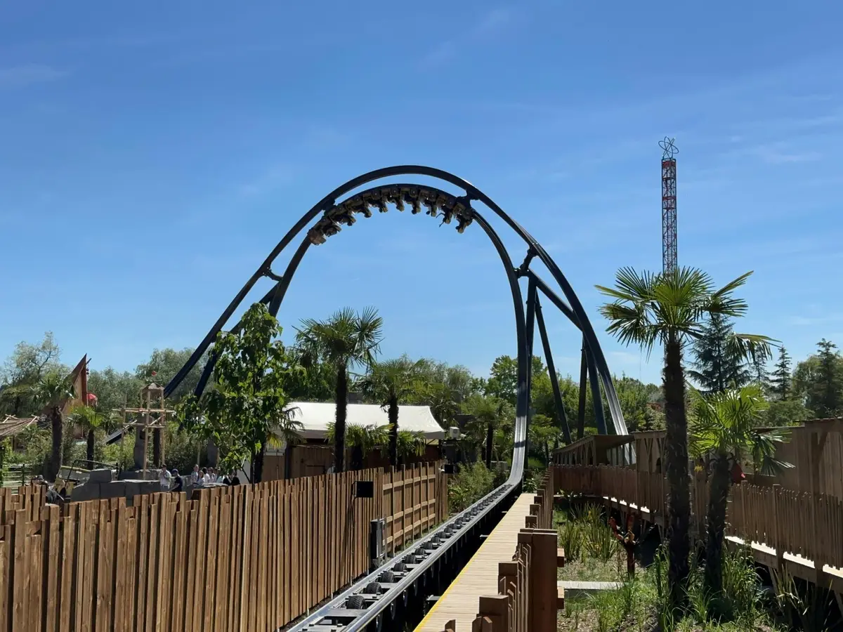 Une montagne russe remplie de passagers suspendus à l'envers au-dessus d'une clôture en bois. Il y a des arbres et un ciel bleu clair.