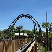 Une montagne russe remplie de passagers suspendus à l'envers au-dessus d'une clôture en bois. Il y a des arbres et un ciel bleu clair. &copy; Walibi Rhone Alpes
