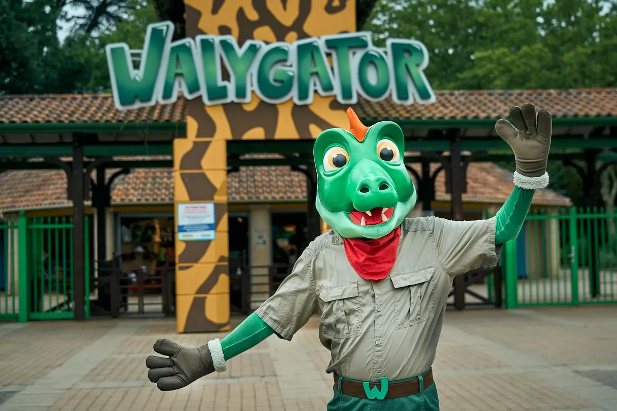Une personne déguisée en dragon s'agite devant l'entrée d'un parc d'attractions où l'on peut lire WALYGATOR.