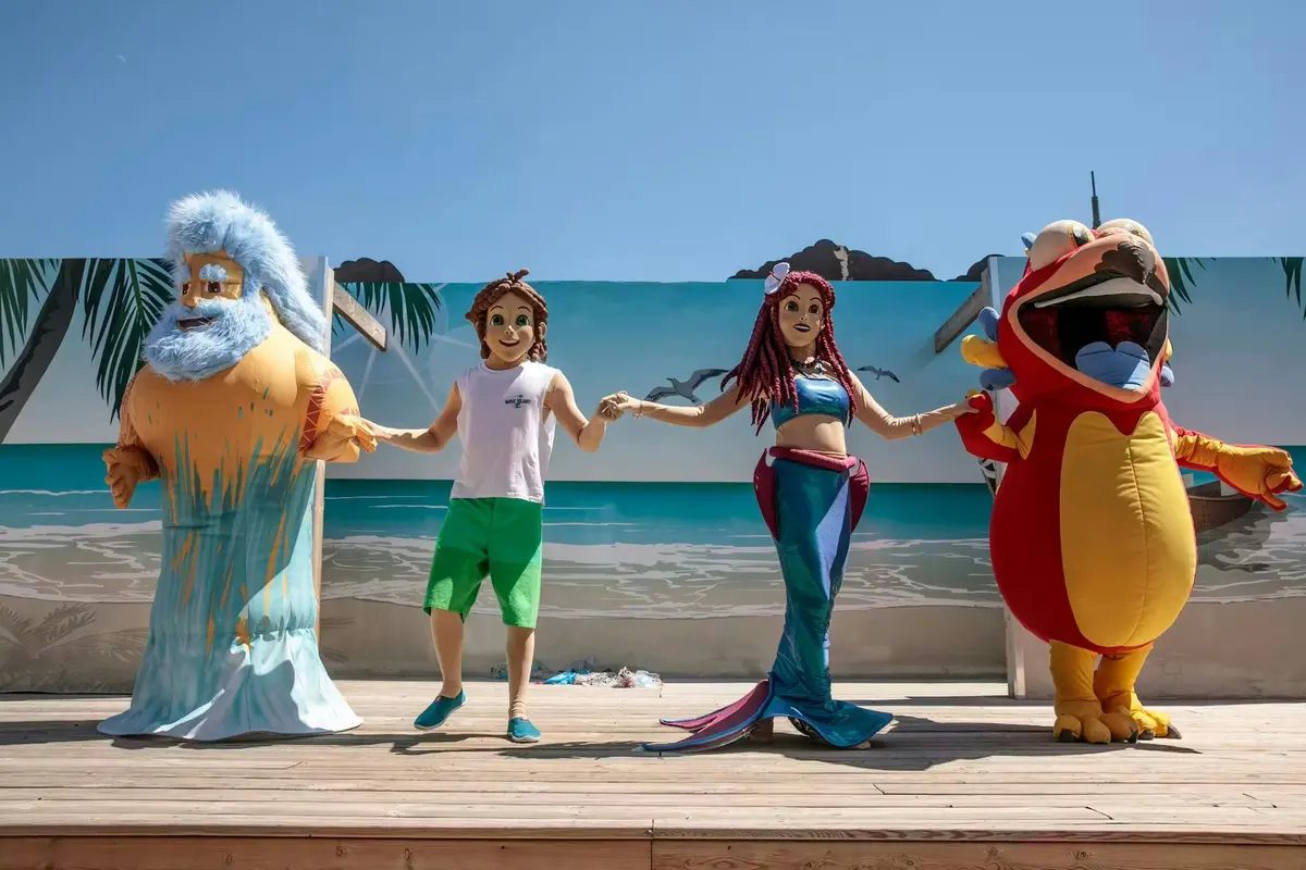 Trois personnes vêtues de costumes colorés, ressemblant à une sirène, un garçon et un homard, se tiennent sur une scène avec une toile de fond sur le thème de la plage.