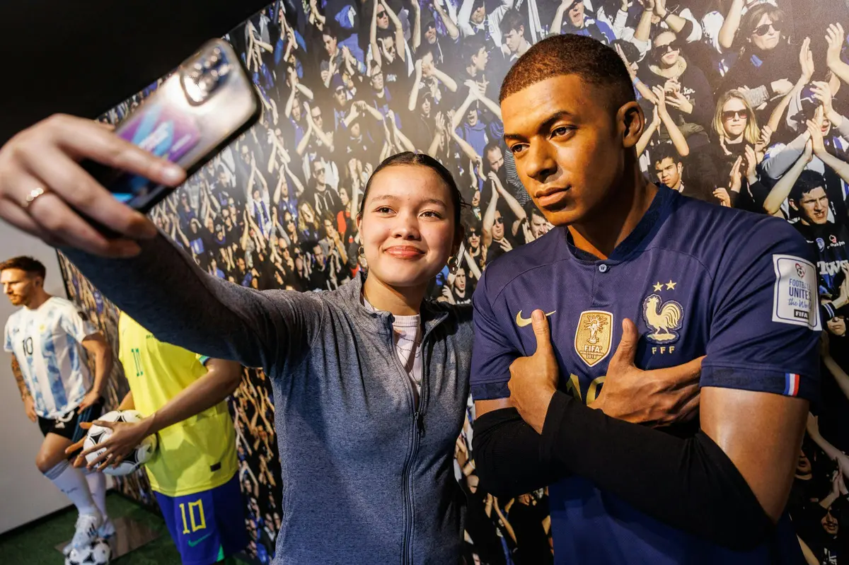 Une personne prend un selfie avec une statue de cire représentant un footballeur portant le maillot de l'équipe de France, devant une toile de fond représentant une foule.