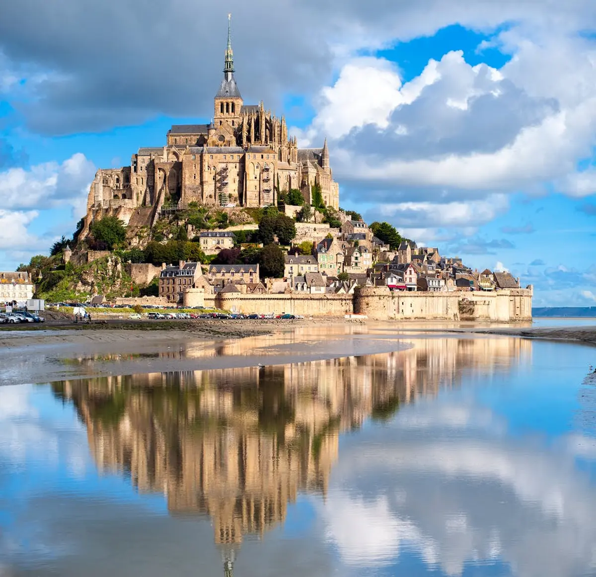 Le Mont Saint-Michel et son abbaye, entourés d'eau et se reflétant dans le calme, sous un ciel bleu partiellement nuageux.