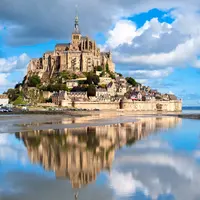 Le Mont Saint-Michel et son abbaye, entourés d'eau et se reflétant dans le calme, sous un ciel bleu partiellement nuageux. DR