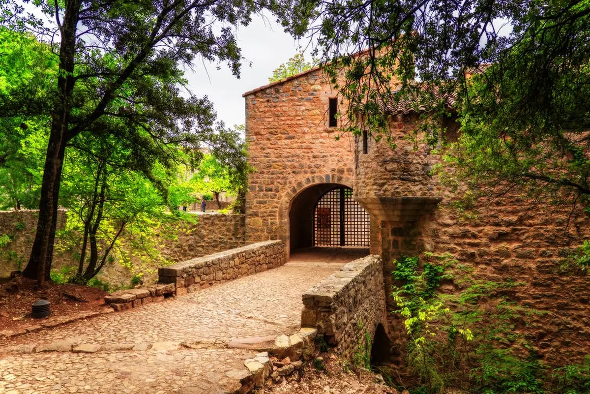 Pont de pierre menant à un portail rustique et arqué, entouré d'arbres et de feuillages verdoyants.