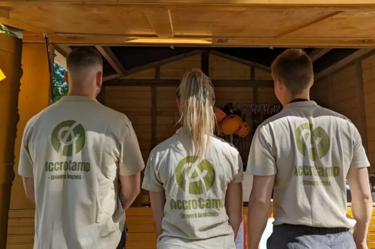 Trois personnes portant le logo AccroCamp sur leur chemise se tiennent face à une structure en bois avec des casques à l'intérieur.