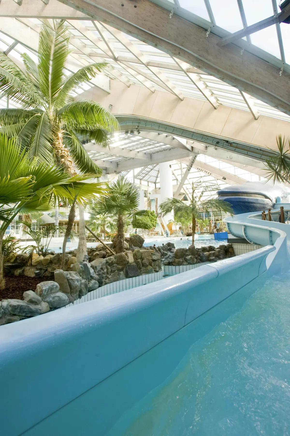 Parc aquatique intérieur sur le thème des tropiques, avec un toboggan bleu, des palmiers, des rochers et une grande verrière.