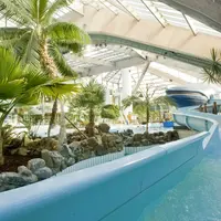 Parc aquatique intérieur sur le thème des tropiques, avec un toboggan bleu, des palmiers, des rochers et une grande verrière. DR