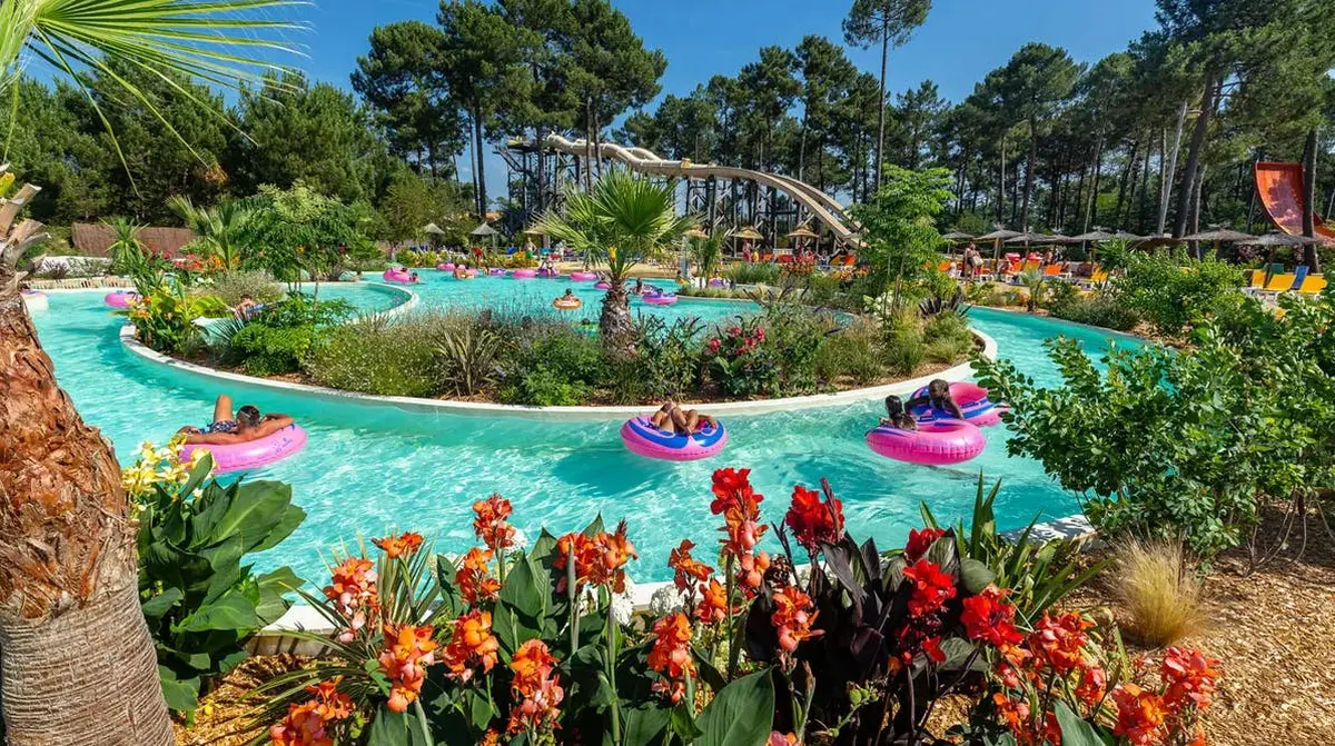Un parc aquatique où des visiteurs flottent sur des bouées dans une rivière tranquille, au milieu d'une végétation luxuriante et de fleurs aux couleurs vives.