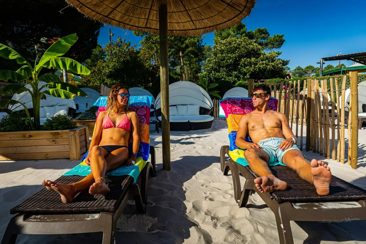 Un homme prend un bain de soleil sur une chaise longue sous un parasol de paille sur une plage de sable, avec une autre personne se relaxant à proximité.