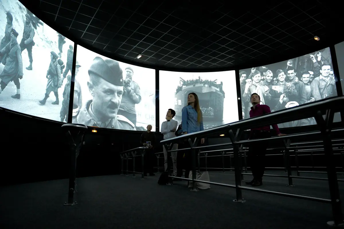 Des personnes regardent des séquences historiques de guerre en noir et blanc sur de grands écrans incurvés dans un espace d'exposition moderne au plafond sombre.