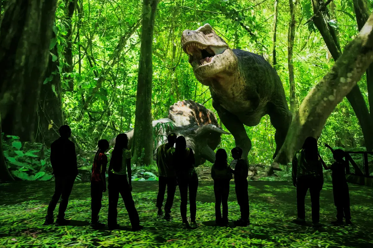 Silhouettes de personnes se tenant devant une scène de jungle vivante avec de grandes projections de dinosaures plus vrais que nature.