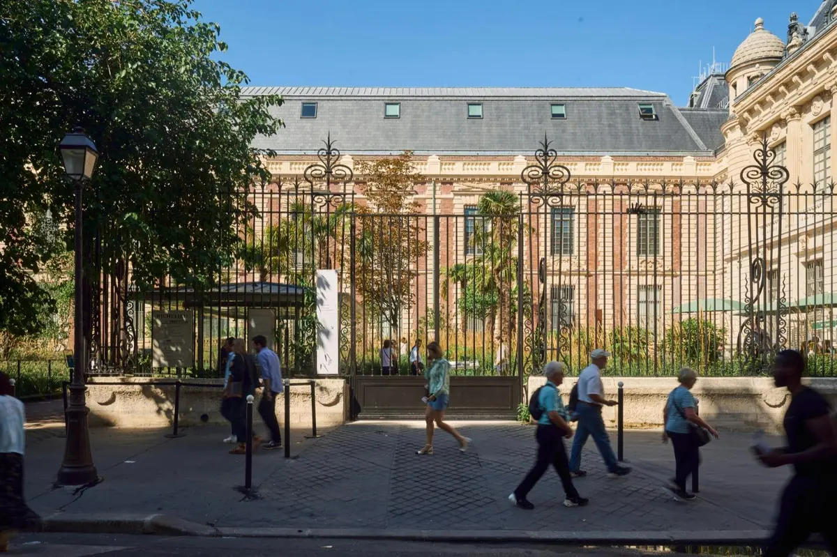 Des passants marchent sur un trottoir devant un immeuble clôturé, doté d'une façade en briques et d'un toit en ardoise, par une journée ensoleillée.